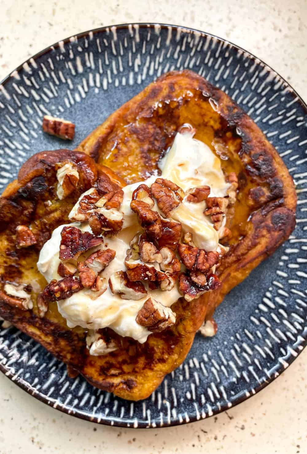 Two slices of golden-brown pumpkin French toast on a plate, topped with fluffy pumpkin whipped cream, chopped candied pecans and sweet maple syrup