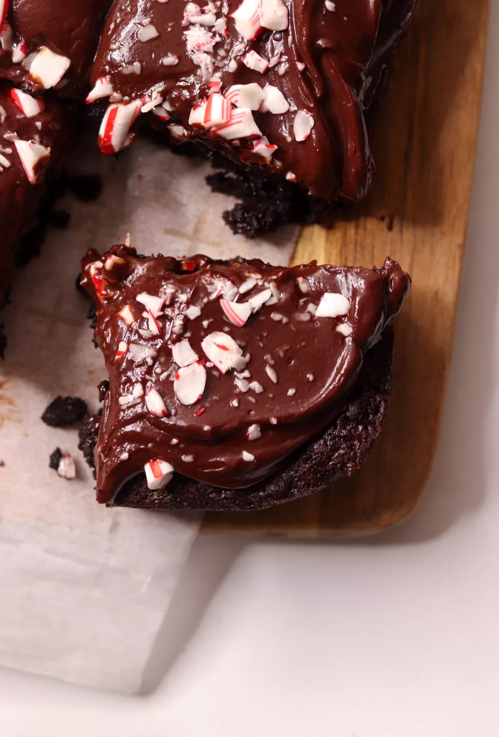 A single peppermint mocha brownie on a wooden cutting board covered with parchment paper. The brownie is pulled away from the rest of the brownies, swirled with dark chocolate ganache and sprinkled with crushed peppermint.