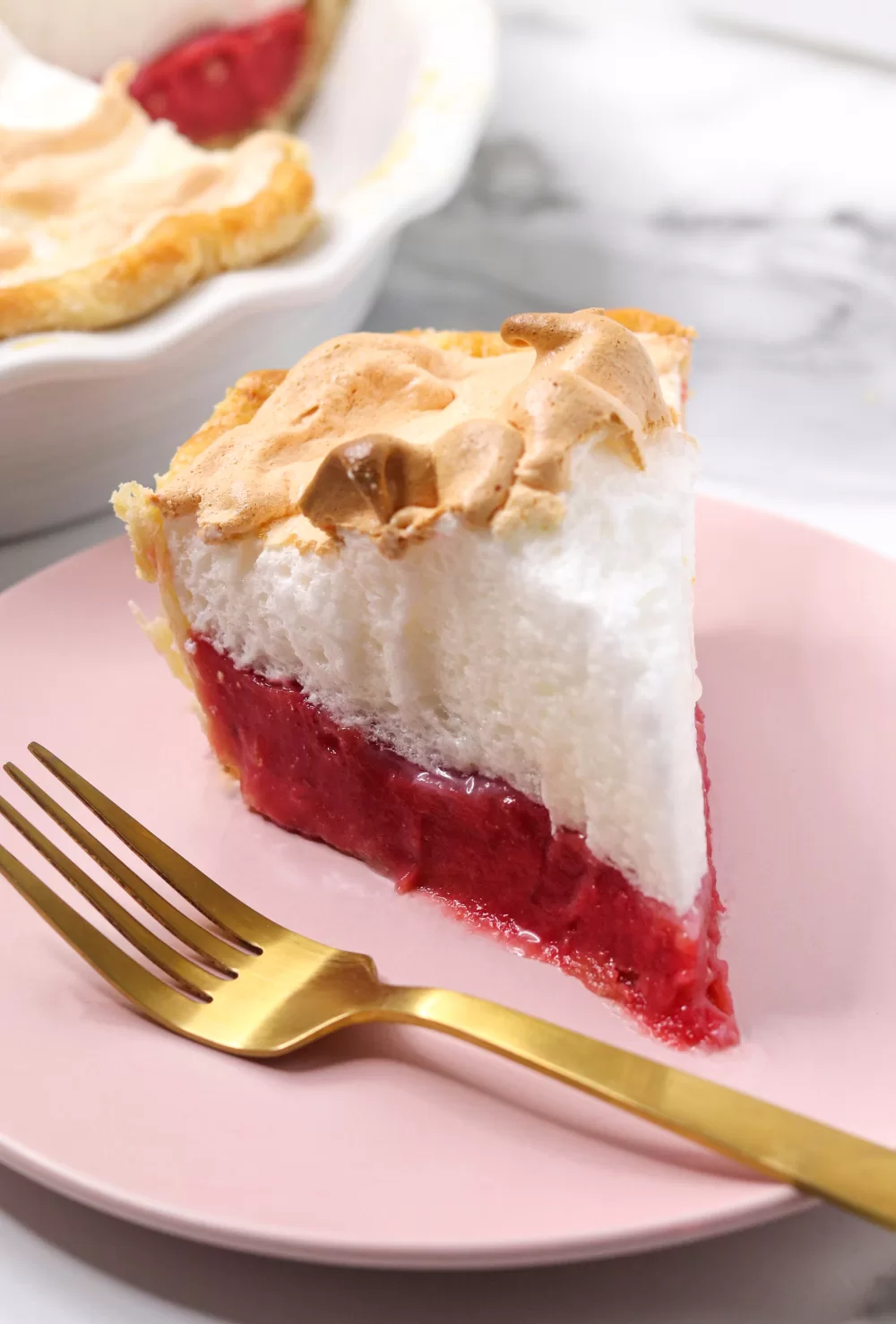 A slice of cranberry meringue pie on a pink plate with a gold fork. It has layers of flaky pie crust, cranberry orange curd and fluffy meringue.