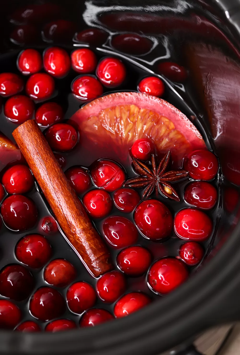 Mulled red wine in a slow cooker, laden with fresh cranberries, a cinnamon stick, star anise and orange slices.
